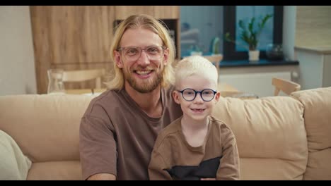 Portrait-of-a-happy-blond-man-in-glasses-and-his-little-albino-son-in-blue-glasses-who-are-sitting-on-a-cream-sofa-in-a-modern-apartment.-A-little-albino-boy-with-white-hair-wearing-blue-glasses-sits-and-rejoices-with-his-father-in-a-modern-apartment-in-front-of-school-in-the-morning