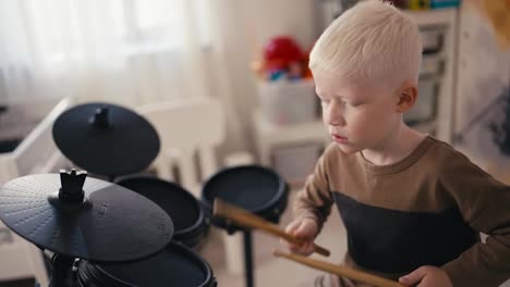 Close-up-shot-of-a-happy-albino-boy-with-white-hair-playing-a-black-electronic-drum-kit-using-special-wooden-sticks-in-his-room-during-a-day-off-developing-his-hobby-and-talent