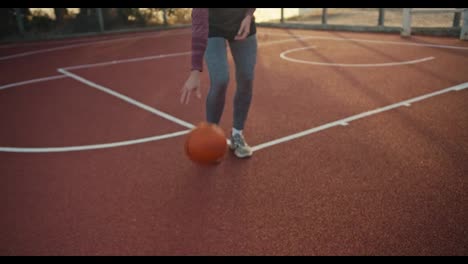 Close-up-a-girl-in-a-sports-uniform-hits-an-orange-basketball-ball-from-a-red-floor-on-a-street-court-at-sunrise-in-summer.-The-girl-goes-in-for-sports-and-develops-the-ability-to-play-basketball-in-the-morning