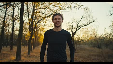 Portrait-of-a-man-with-curly-hair-in-a-black-sports-uniform-who-looks-confidently-at-the-camera-and-smiles-while-holding-his-arms-crossed-on-his-chest-In-the-autumn-forest-in-the-morning