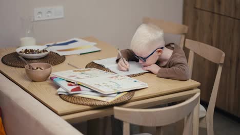 Ein-Kleiner-Albino-Junge-Mit-Weißem-Haar-Und-Blauer-Brille-Schreibt-Mit-Einem-Stift-In-Sein-Notizbuch-Und-Bereitet-Seine-Hausaufgaben-Vor-Dem-Nächsten-Schultag-In-Einer-Modernen-Wohnung-In-Der-Küche-Vor