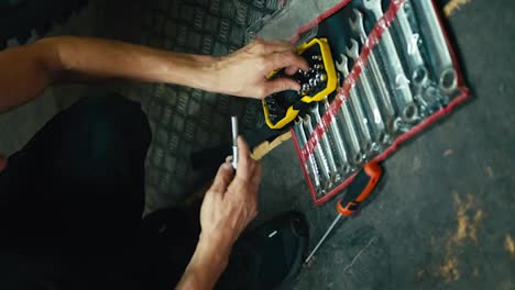 Vertical-video:-close-up:-a-male-mechanic-with-his-sinewy-hands-selects-the-tool-he-needs-among-a-set-of-tools-on-the-floor-in-the-workshop