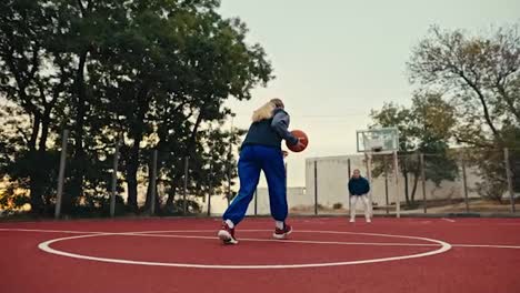 Shot-from-below-a-blonde-girl-in-a-sports-uniform-runs-along-a-red-street-basketball-court-and-throws-the-ball-into-a-basketball-hoop-in-the-morning