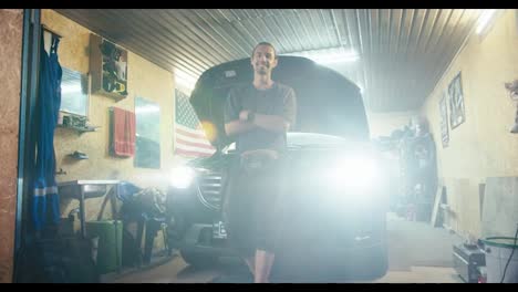 Portrait-of-a-confident-motorist-with-collected-hair-in-a-gray-T-shirt-who-looks-at-the-camera-and-leans-on-the-hood-of-his-car-in-his-garage-workshop