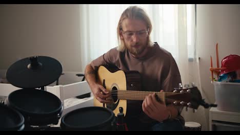 Un-Hombre-Rubio-Con-Gafas-Y-Barba-Con-Una-Camiseta-Marrón-Toca-Una-Guitarra-Acústica-Amarilla.-Cerca-De-él-Hay-Una-Batería.-Desarrollo-De-Talentos-Y-Aficiones-En-Casa.