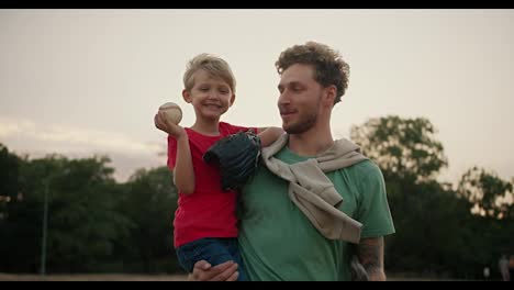 Happy-little-blond-boy-in-a-red-T-shirt-holds-a-baseball-in-his-hands-and-throws-it-into-the-glove-of-his-dad-in-a-Green-T-shirt.-Portrait-of-a-boy-and-his-dad-playing-baseball-in-the-park