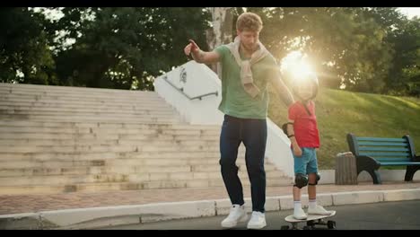 A-father-in-a-green-t-shirt-with-curly-hair-and-blue-jeans-helps-his-little-son-in-a-red-t-shirt-and-a-protective-black-helmet-ride-a-skateboard-in-the-park-on-the-road.-Happy-dad-and-son-spending-time-in-the-park