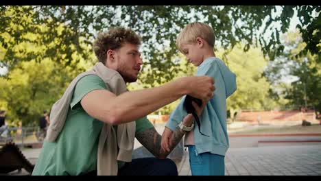 A-man-with-curly-hair-and-stubble-in-a-green-T-shirt-helps-his-son-in-a-blue-sweater-put-on-his-elbow-pads-before-riding-a-skateboard-at-a-skatepark-in-the-park.-Little-blond-boy-preparing-to-ride-a-skateboard