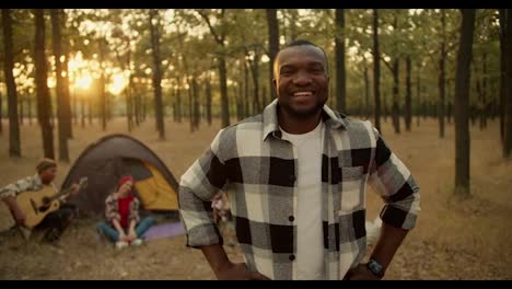 Portrait-of-a-Happy-man-with-black-skin-in-a-checkered-shirt-who-stands-straight-and-poses-against-the-background-of-the-rest-of-the-group-of-hikers-and-tents-in-a-Sunny-summer-green-forest