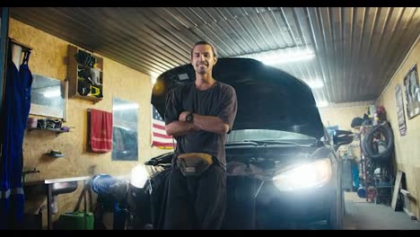 Portrait-of-a-confident-motorist-mechanic-who-puts-his-hands-on-his-chest-and-looks-at-the-camera-and-smiles-while-leaning-on-the-hood-of-his-car-in-a-workshop-garage-in-the-evening