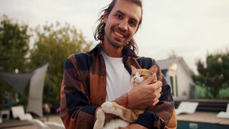 Portrait:-A-brunette-guy-with-long-hair-holds-a-red-cat-and-strokes-it.-Rest-in-the-country-house