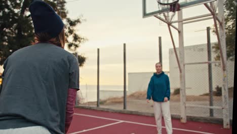 Shot-from-behind-a-blonde-girl-with-a-bob-hairstyle-receives-a-basketball,-catches-it-and-throws-it-into-the-basketball-hoop-while-playing-with-her-friend-on-a-summer-morning