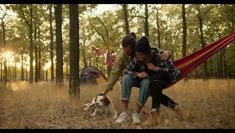 A-happy-couple,-a-brunette-man-in-a-light-green-jacket-and-a-blonde-girl-in-a-black-hat,-are-sitting-on-a-red-hammock-and-petting-their-white-brown-dog-against-the-background-of-a-gray-tent-in-a-sunny-summer-green-forest