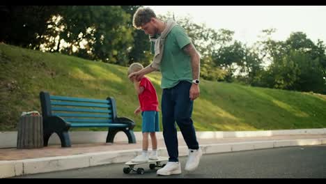 A-father-in-a-green-T-shirt-and-blue-jeans-helps-his-son-in-a-red-T-shirt-and-blue-shorts-ride-a-skateboard-along-the-road-in-the-park.-Happy-father-and-son-spending-time-active-leisure