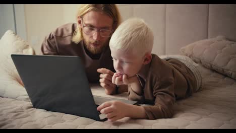 Un-Hombre-Rubio-Con-Gafas-Y-Barba,-Junto-Con-Su-Pequeño-Hijo-Albino-De-Ojos-Azules-Con-Una-Chaqueta-Marrón,-Miran-Una-Película-En-Una-Computadora-Portátil-Y-Dibujos-Animados-Educativos-Durante-Su-Relajación-Y-Pasan-Tiempo-Juntos.