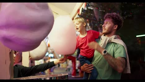 A-little-blond-boy-in-a-red-T-shirt-takes-pink-cotton-candy-from-a-sweet-shop-in-an-amusement-park-with-his-dad.-A-little-boy-in-a-red-T-shirt-sits-in-the-arms-of-his-dad-in-a-Green-T-shirt-and-eats-cotton-candy