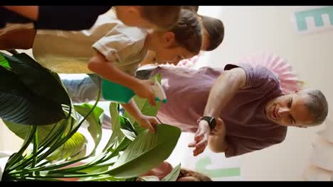 Vertical-video-of-a-man-with-gray-hair-in-a-purple-T-shirt-teaching-preschool-children-how-to-properly-water-flowers-using-a-spray-bottle-in-a-club-room-to-prepare-children-for-school