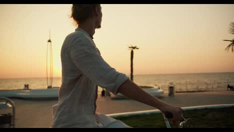 A-happy-young-guy-in-a-light-shirt-rides-a-bicycle-along-the-beach-where-palm-trees-grow-near-the-sea-at-sunrise-in-summer.-A-guy-does-morning-exercises-and-drives-along-the-beach-in-the-summer