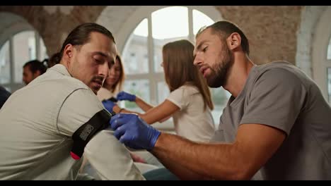 Close-up-shooting:-A-bearded-brunette-man-in-a-gray-T-shirt-puts-a-tourniquet-on-the-hand-of-a-brunette-man-with-long-hair-during-a-practical-medical-course