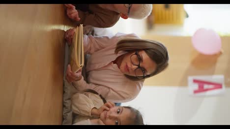 Vertical-video-of-a-woman-with-a-bob-hairstyle-in-glasses-in-a-pink-shirt-reading-a-book-to-children-in-a-special-club-for-preparing-children-for-school-and-telling-her-and-teaching-them