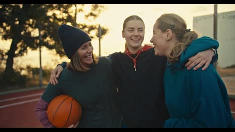 Close-up-a-trio-of-blonde-girls-in-sportswear-with-a-basketball-on-a-red-summer-court-on-the-street-stand-hugging-and-talking-after-their-match-in-the-morning-at-sunrise