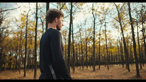 A-serious-man-with-curly-hair-with-a-beard-in-a-black-sports-uniform-looks-at-his-watch,-notes-the-time-and-starts-his-jog-in-the-autumn-forest-in-the-morning