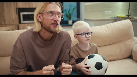 A-happy-blond-man-with-a-beard-and-glasses,-together-with-his-little-albino-son,-are-rooting-for-their-favorite-football-team-and-watching-the-match.-The-little-albino-boy-is-holding-a-soccer-ball-in-his-hands-and-then-throwing-it-up.-After-the-happiness-of-Victory