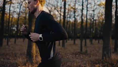 Un-Atleta-Masculino-Con-Uniforme-Deportivo-Negro-Y-Cabello-Rizado-Mira-Su-Reloj-Y-Luego-Continúa-Su-Carrera-Mientras-Trota-Por-La-Mañana-En-El-Bosque-Otoñal-Soleado.