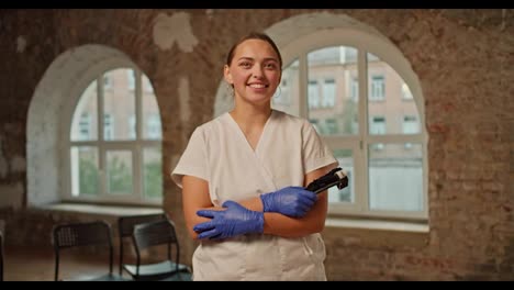 Retrato:-Una-Doctora-Profesional-Con-Un-Uniforme-Médico-Blanco-Se-Encuentra-Frente-A-Un-Fondo-De-Paredes-De-Ladrillo-Y-Grandes-Ventanas-Blancas.