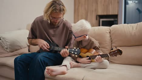 Ein-Kleiner-Albino-Junge-Mit-Weißen-Haaren-Und-Blauer-Brille-Lernt-Zusammen-Mit-Seinem-Vater-Und-Einem-Blonden-Mann-Mit-Brille-Und-Bart-Das-Ukulele-Spielen,-Während-Er-Abends-Auf-Einem-Cremefarbenen-Großen-Sofa-In-Einer-Modernen-Wohnung-Sitzt