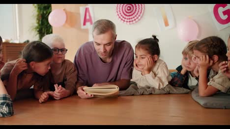 A-man-with-gray-hair-in-a-purple-T-shirt,-a-teacher-reads-a-book-to-his-students-who-lie-on-the-floor-on-special-pillows-and-listen-carefully-to-their-teacher-in-a-club-for-preparing-children-for-school