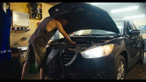 A-male-master-mechanic-with-a-short-haircut-in-a-gray-T-shirt-and-green-shorts-repairs-the-hood-of-a-car-with-the-headlights-on-and-a-slight-haze-in-his-garage-workshop