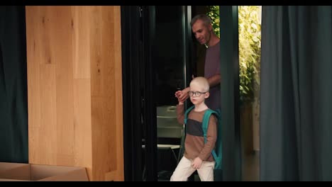 Ein-Vater-Mit-Grauen-Haaren-In-Einem-Lila-T-Shirt-Kam-Mit-Seinem-Sohn-Mit-Weißen-Haaren,-Einer-Runden-Brille-Und-Einem-Grünen-Rucksack-Zu-Einem-Club-Zur-Vorbereitung-Der-Kinder-Auf-Die-Schule,-Wo-Sie-Von-Einer-Lehrerin-In-Einem-Weißen-Hemd-Empfangen-Wurden