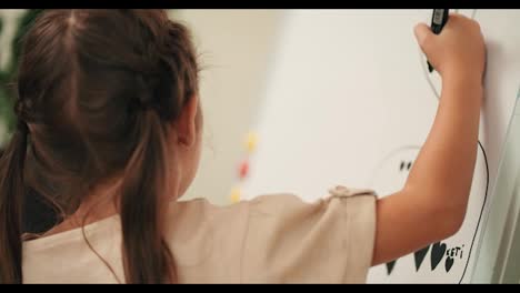 A-girl-using-a-black-marker-draws-on-a-large-white-sheet-of-paper-that-stands-on-an-easel-in-a-drawing-mug-in-the-room.-The-preschool-girl-draws-with-a-large-marker