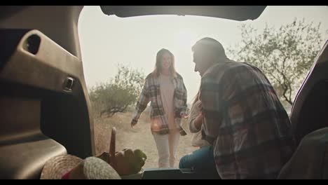 A-happy-man-with-gray-hair-in-a-checkered-shirt-meets-his-wife,-a-brunette-girl-in-a-green-checkered-shirt,-and-their-little-daughter-running-towards-him-while-the-man-sits-in-the-open-trunk-of-a-car-and-looks-at-his-loved-ones-during-a-picnic-in-the-summer