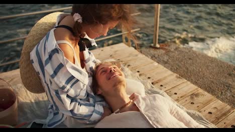 A-happy-blond-guy-with-stubble-in-light-clothes-lies-on-the-lap-of-his-brown-haired-girlfriend-at-a-picnic-on-the-pier-near-the-sea.-A-guy-and-a-girl-relax-near-the-sea