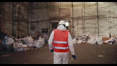 Rear-view-of-a-man-in-a-red-safety-vest-and-white-uniform-walking-through-a-tall-waste-recycling-plant.-A-huge-room-with-polar-bears-with-garbage-that-will-be-recycled-in-a-huge-plant