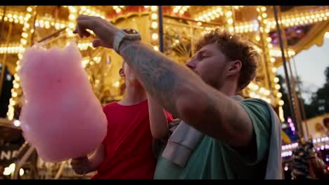 A-little-blond-boy-in-a-red-T-shirt-eats-pink-cotton-candy-with-his-dad-in-a-Green-T-shirt-against-the-backdrop-of-a-beautiful-bright-attraction-with-yellow-flowers-in-an-amusement-park-in-the-evening