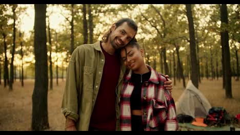 A-happy-couple,-a-brunette-man-in-a-green-shirt-and-a-Black-skinned-girl-in-a-checkered-shirt-stand-and-look-at-the-camera-and-smile-against-the-background-of-the-accessories-they-took-with-them-on-a-hike-in-a-green-Sunny-summer-forest