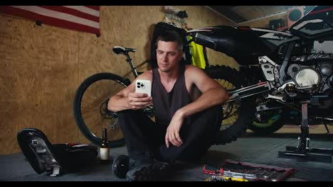 Male-mechanic-with-a-short-haircut-in-a-gray-T-shirt-works-on-his-white-phone-in-the-workshop-during-a-break-while-repairing-a-motorcycle.-A-man-sits-near-the-tools-in-his-workshop