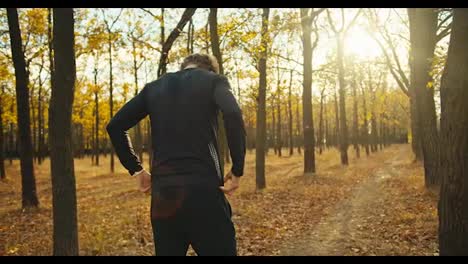 From-behind-a-happy-man-with-curly-hair-in-black-sports-autumn-clothes-in-an-autumn-forest-with-fallen-leaves-rests-after-his-jog-and-prepares-for-new-sports-activities-on-a-sunny-day-in-autumn