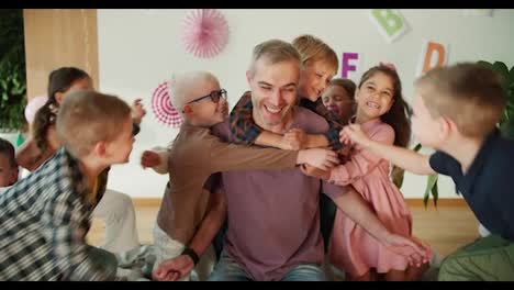 Eine-Gruppe-Von-Kindern-Umarmt-Ihren-Lehrer,-Einen-Mann-Mit-Grauen-Haaren-In-Einem-Lila-T-Shirt,-Bei-Ihrer-Ersten-Unterrichtsstunde-Zur-Vorbereitung-Der-Kinder-Auf-Die-Schule-In-Einem-Speziellen-Club.-Die-Glücklichen-Kinder-Lieben-Ihren-Lehrer-Und-Umarmen-Ihn