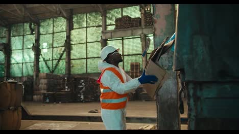 A-man-with-Black-skin-in-a-white-uniform-and-an-orange-vest-with-a-white-helmet-picks-up-waste-paper-that-a-colleague-gives-him-from-the-trunk-of-a-truck-during-unloading-at-a-waste-processing-plant