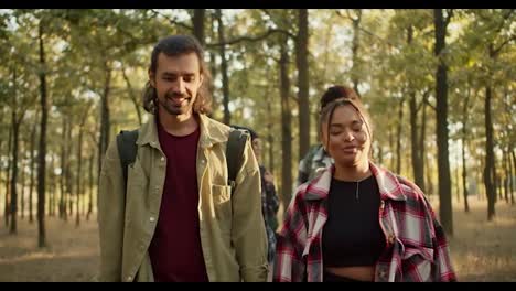 Happy-group-on-a-hike-front-view-man-and-girl-in-hiking-clothes-walking-along-the-summer-forest.-A-girl-with-Black-skin-in-a-checkered-shirt-walks-through-the-forest-and-communicates-with-a-brunette-man-with-stubble-in-a-light-green-jacket