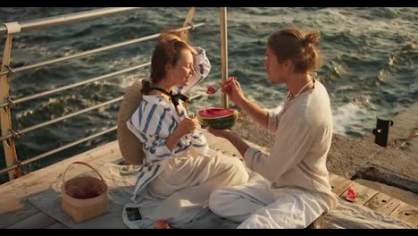 A-blond-guy-with-stubble-in-light-clothes-and-a-brown-haired-girl-with-a-black-bow-and-a-white-and-blue-shirt-are-sitting-on-a-picnic-on-the-pier-near-the-sea-and-eating-watermelon