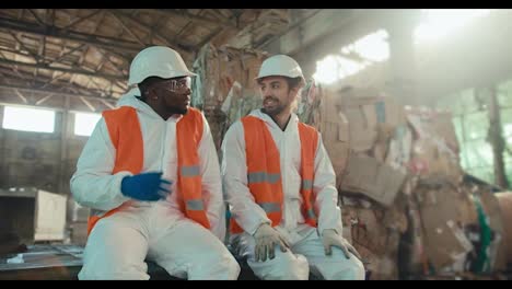 Un-Hombre-Negro-Con-Uniforme-Blanco-Y-Chaleco-Naranja-Se-Sienta-Con-Su-Colega-Moreno,-Con-Barba-Y-Casco-Blanco,-Y-Conversan-Durante-Un-Descanso-En-Una-Gran-Planta-De-Reciclaje-De-Residuos-Y-Papel.