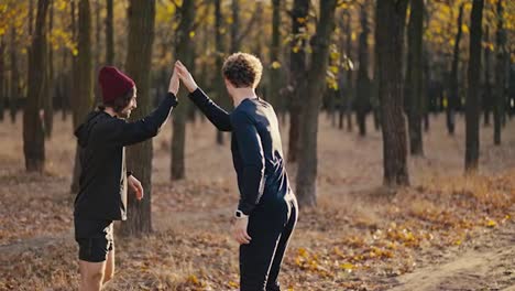 Two-brunette-black-guys-in-sportswear-give-each-other-a-high-five-and-begin-their-jog-in-a-sunny-autumn-forest-along-an-earthen-road