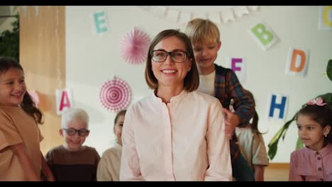 a-group-of-children-hug-their-teacher,-a-blonde-woman-with-a-bob-hairstyle-in-glasses-in-a-white-shirt-at-their-first-lesson-in-a-club-for-preparing-children-for-school-classes.-The-children-love-their-woman-teacher-and-hug-her-during-a-preschool-lesson