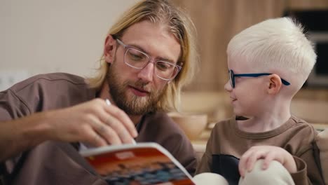 Close-up:-a-blond-man-with-a-beard-and-glasses-helps-his-little-albino-son-with-white-hair-in-blue-round-glasses-to-do-his-homework-and-reads-for-him-what-is-written-in-a-notebook.-Sitting-on-the-sofa-in-a-modern-apartment-in-the-evening