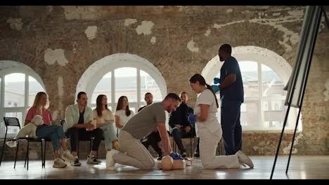 A-bearded-brunette-man-in-a-gray-T-shirt-makes-artificial-respiration-on-a-mannequin,-a-nurse-girl-in-a-white-uniform,-together-with-a-Black-man,-a-doctor-in-a-blue-uniform,-explains-the-rules-for-first-aid-in-front-of-the-public-against-the-background-of-brick-walls-and-large-windows